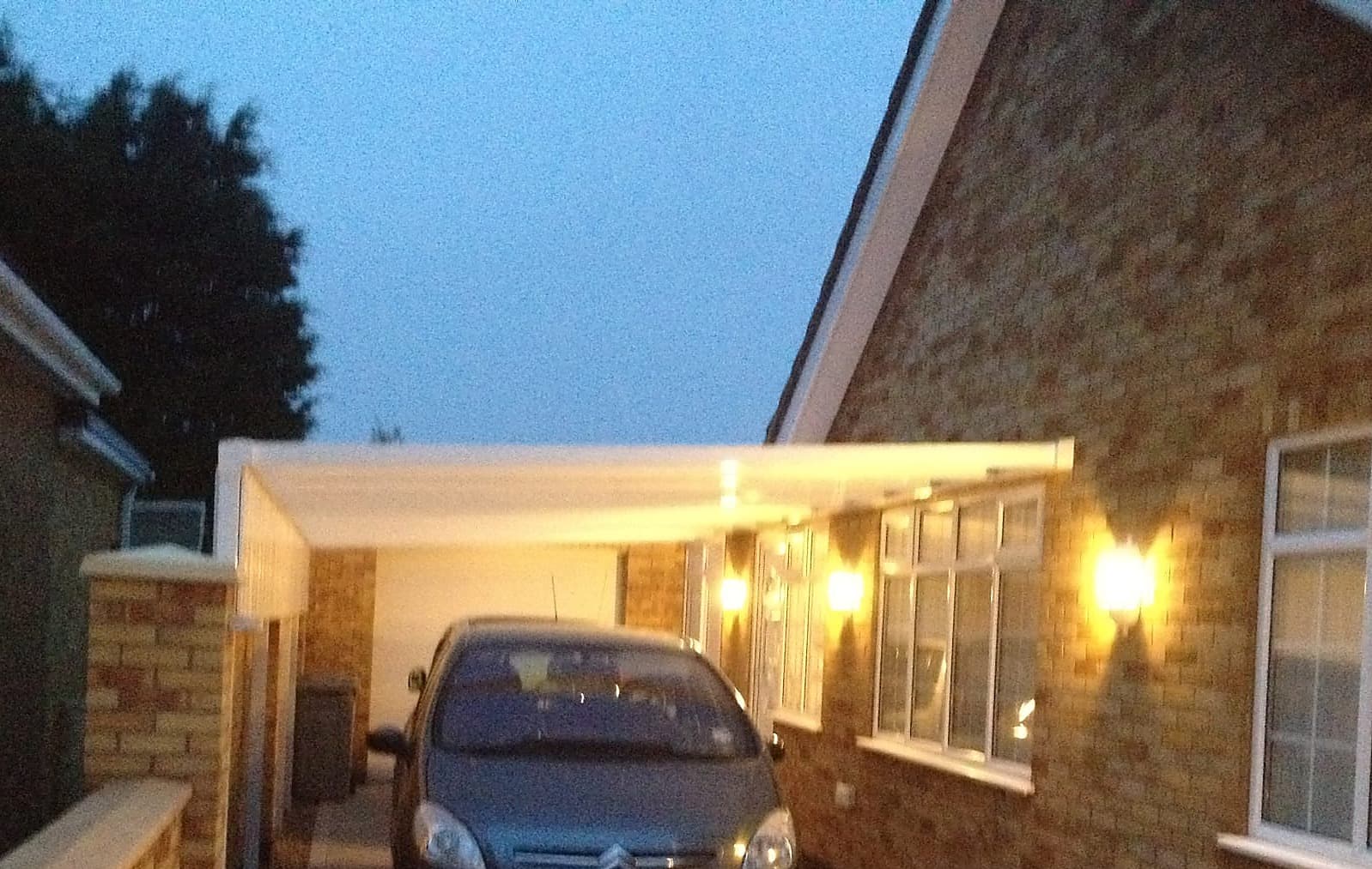 Car parked under a white carport canopy at dusk with the wall lights on