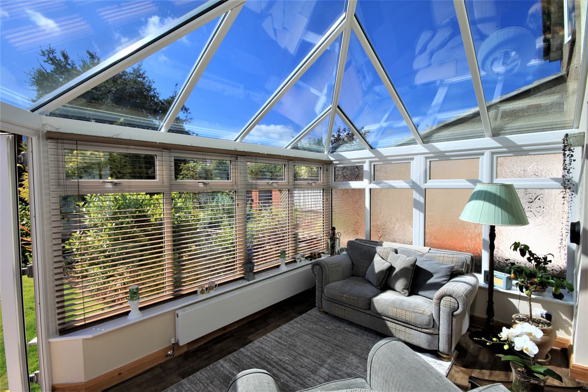 Conservatory interior with a pitched glazed roof, slatted blinds and grey sofas