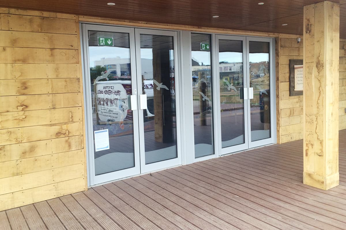 Light grey framed glazed entrance doors in a timber clad building with decking