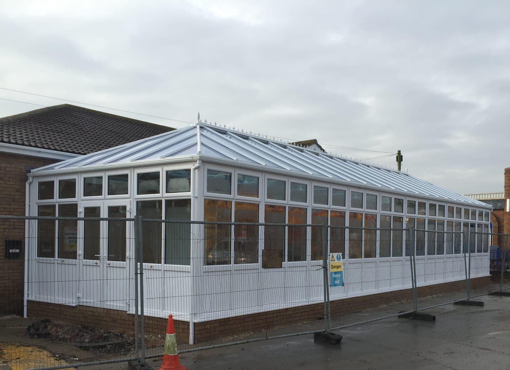 Long white conservatory with a glazed roof and a brick base behind temporary site fencing